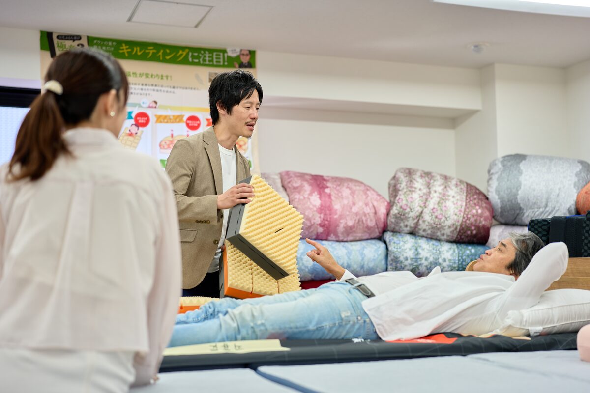 販売会場で、マットレスの断面見本を手にしたスタッフがお客さまにご説明している様子。奥では別のお客さまがマットレスに横になってお試し中